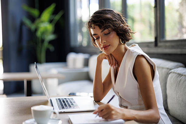 Young businesswoman working with laptop and writing notes at modern cafe